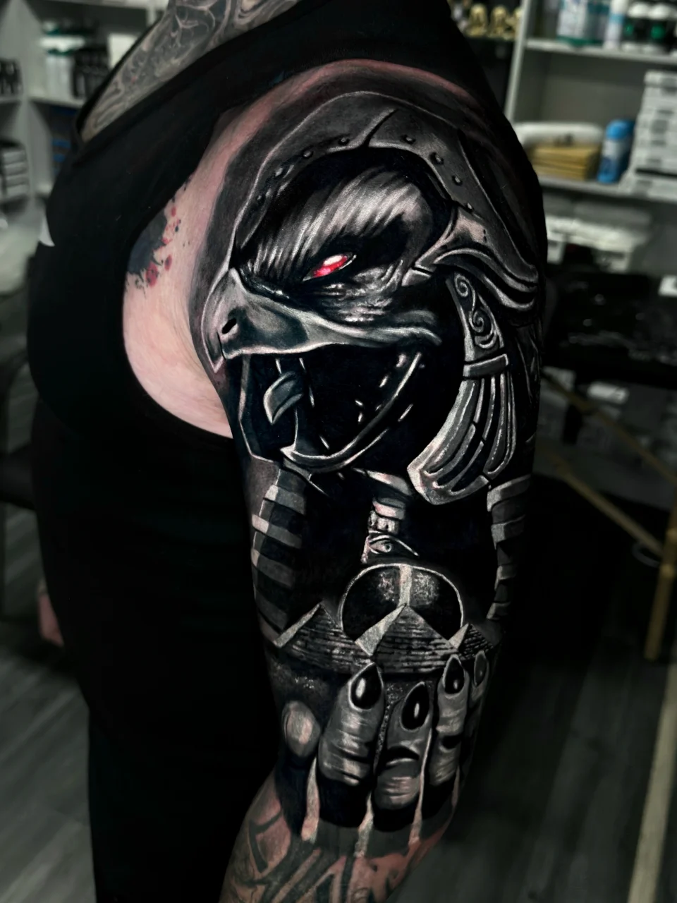 A detailed black and gray tattoo of an Egyptian god with a jackal head and glowing red eyes covers a persons upper arm and shoulder, featuring bold shading and intricate armor-like designs.