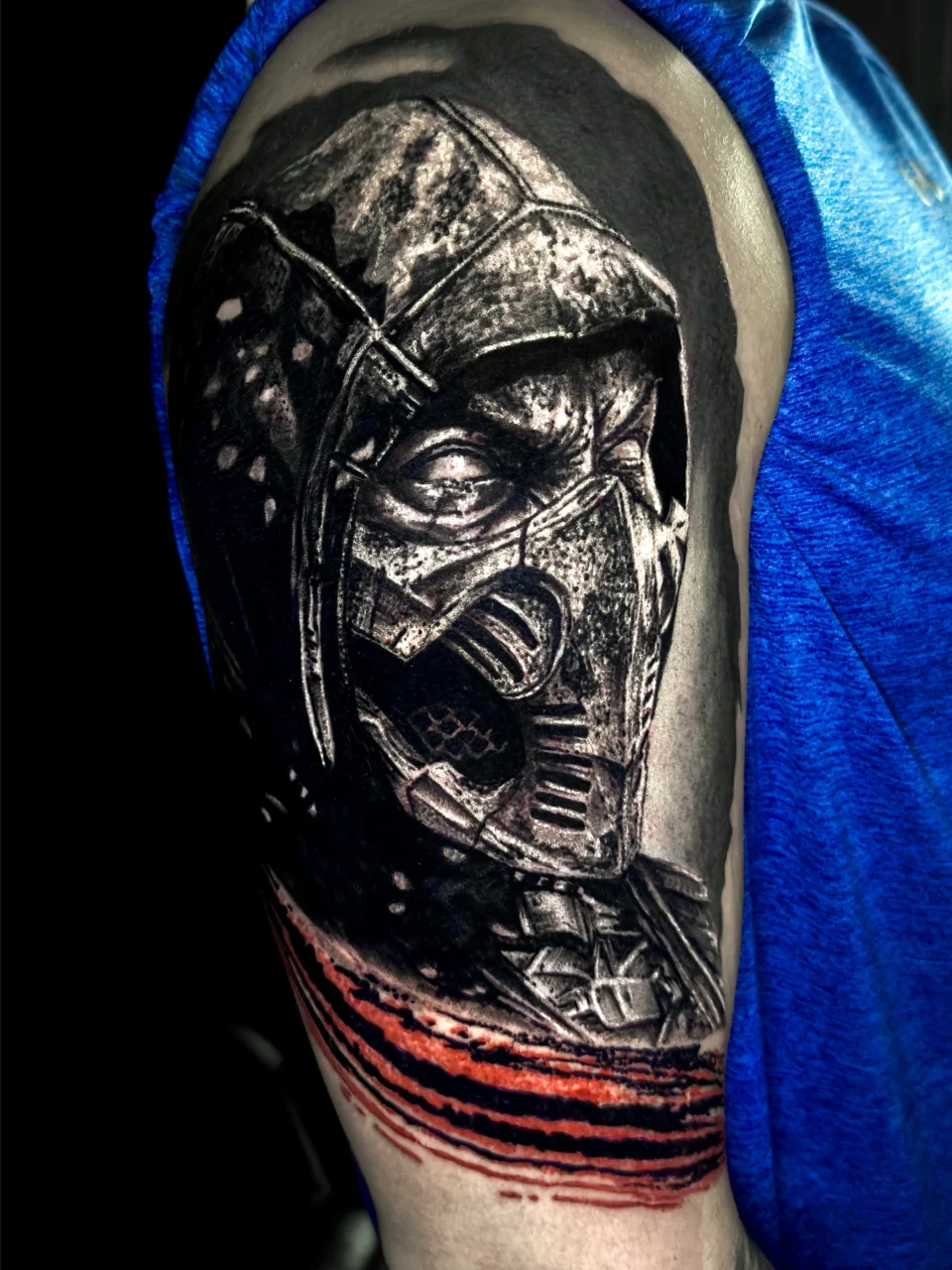 A realistic black and grey tattoo of a masked warrior wearing a hood, inspired by a video game character, is inked on an upper arm. The design includes bold red slashes beneath the portrait.