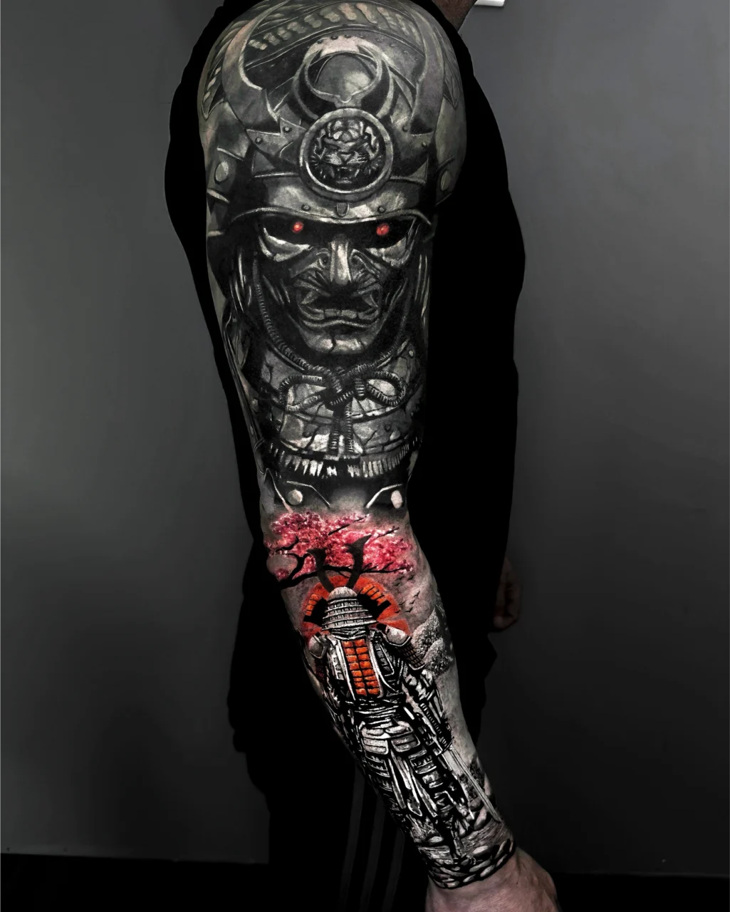 A person’s arm with a detailed sleeve tattoo featuring a samurai helmet and face with glowing red eyes on the upper arm, and a samurai figure standing under pink cherry blossoms on the forearm.