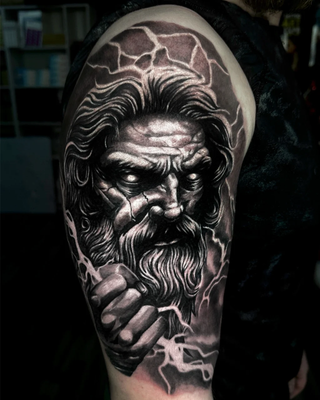 Detailed black and grey tattoo on an upper arm, depicting a fierce bearded mans face with intense eyes, surrounded by lightning and holding a fist, suggesting a mythological or god-like figure.