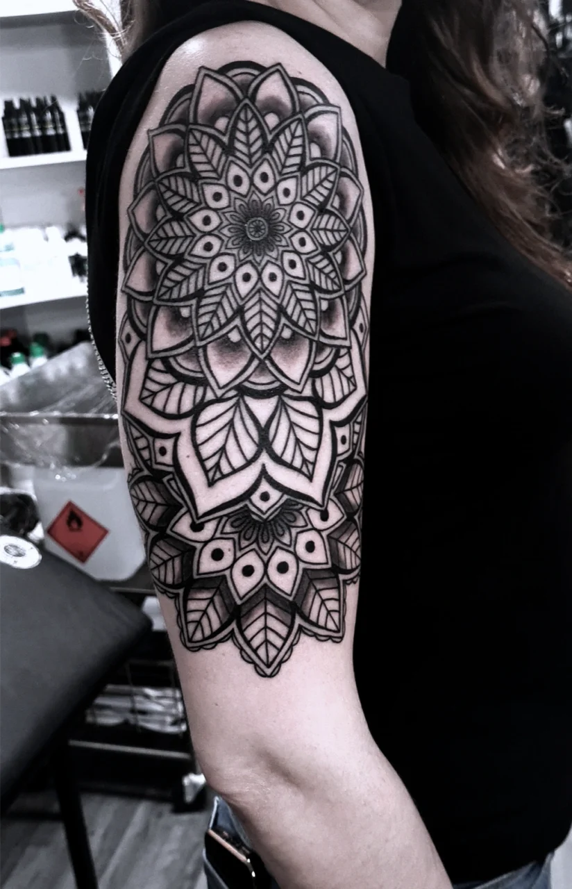 A person with long hair wears a black sleeveless top, displaying a large, intricate mandala tattoo in black ink covering the upper arm and shoulder. Shelves and tattoo equipment are visible in the background.