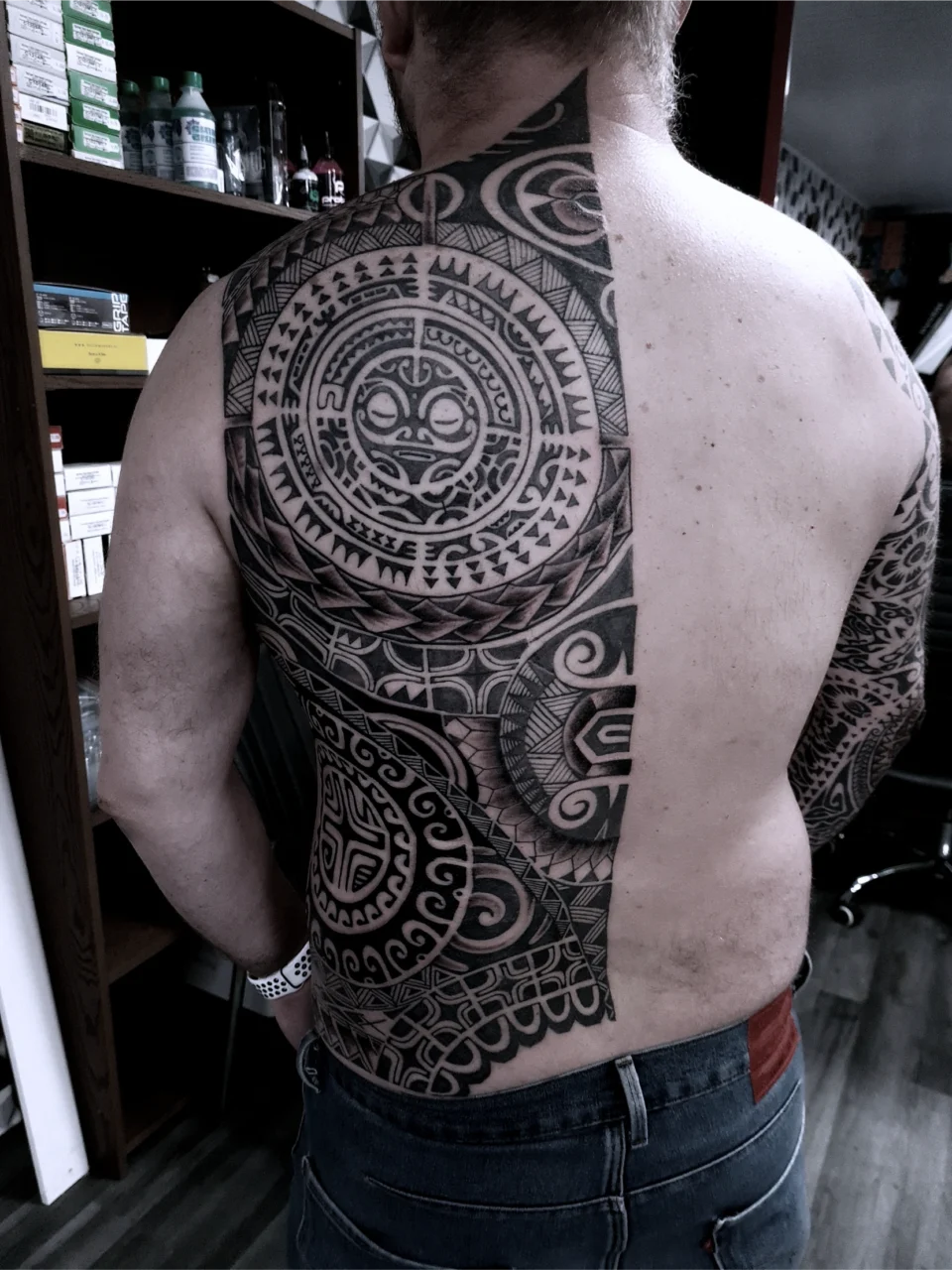 A person with their back exposed displays an intricate, large black tribal tattoo covering half their back, shoulder, and upper arm, contrasting with their untattooed skin on the other side. The tattoo features geometric and circular patterns.
