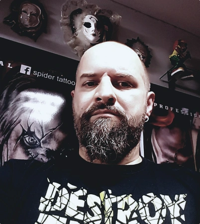 A bearded man with a shaved head takes a selfie in a room decorated with masks and posters, including a large clown face. He wears a black shirt with bold white and yellow text at Spider Ink, a tattoo studio in Dublin.