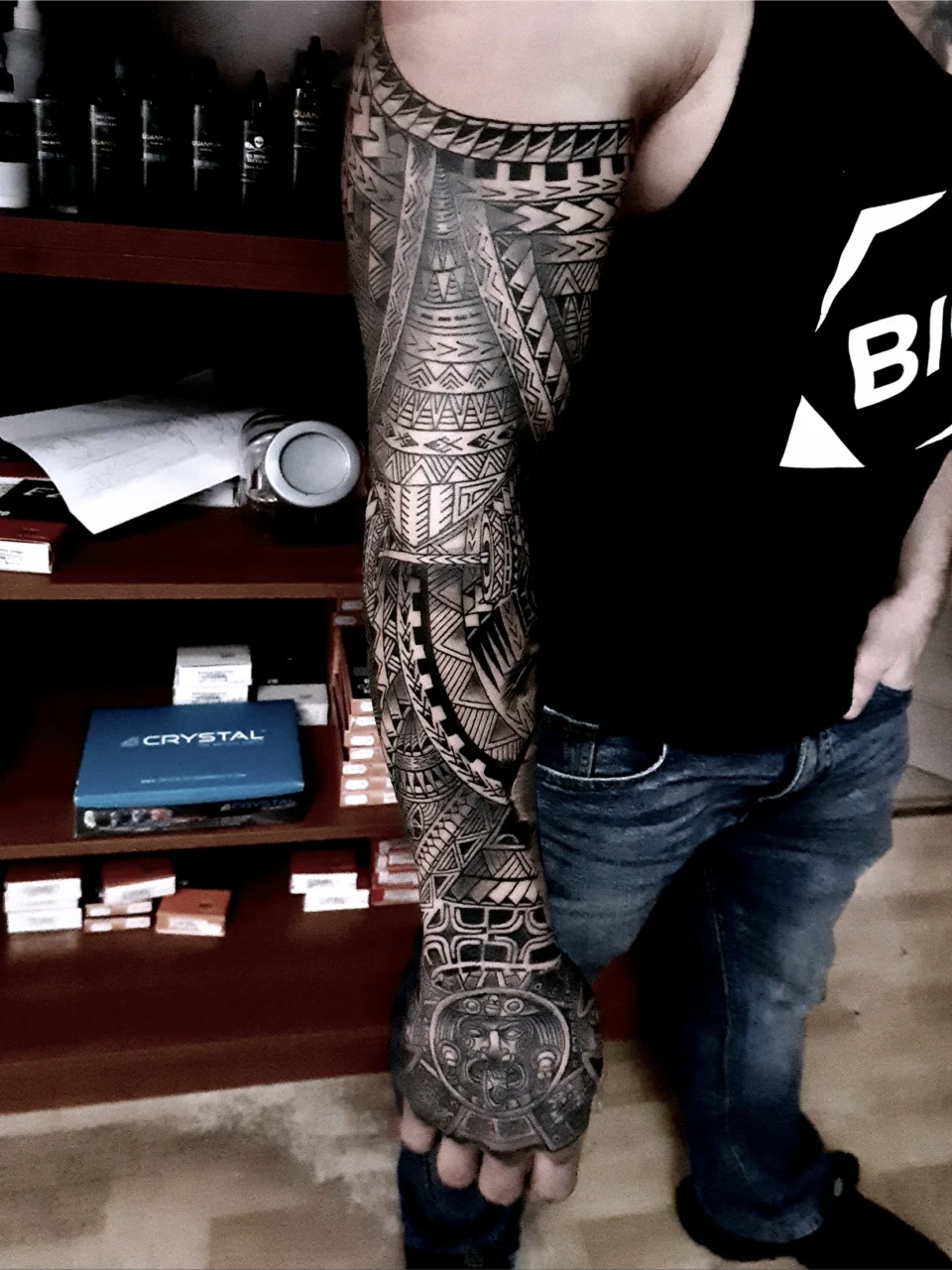 A person with a full-sleeve Polynesian tribal tattoo on their right arm stands in front of a shelf, wearing a sleeveless black shirt and blue jeans. The intricate tattoo features geometric patterns and symbols.