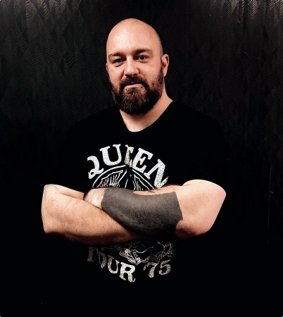 A bearded man with a shaved head stands with arms crossed, wearing a black Queen Tour 75 t-shirt. His large black tattoo, crafted at a top tattoo studio in Dublin, covers his left forearm as he poses against a dark, textured background.