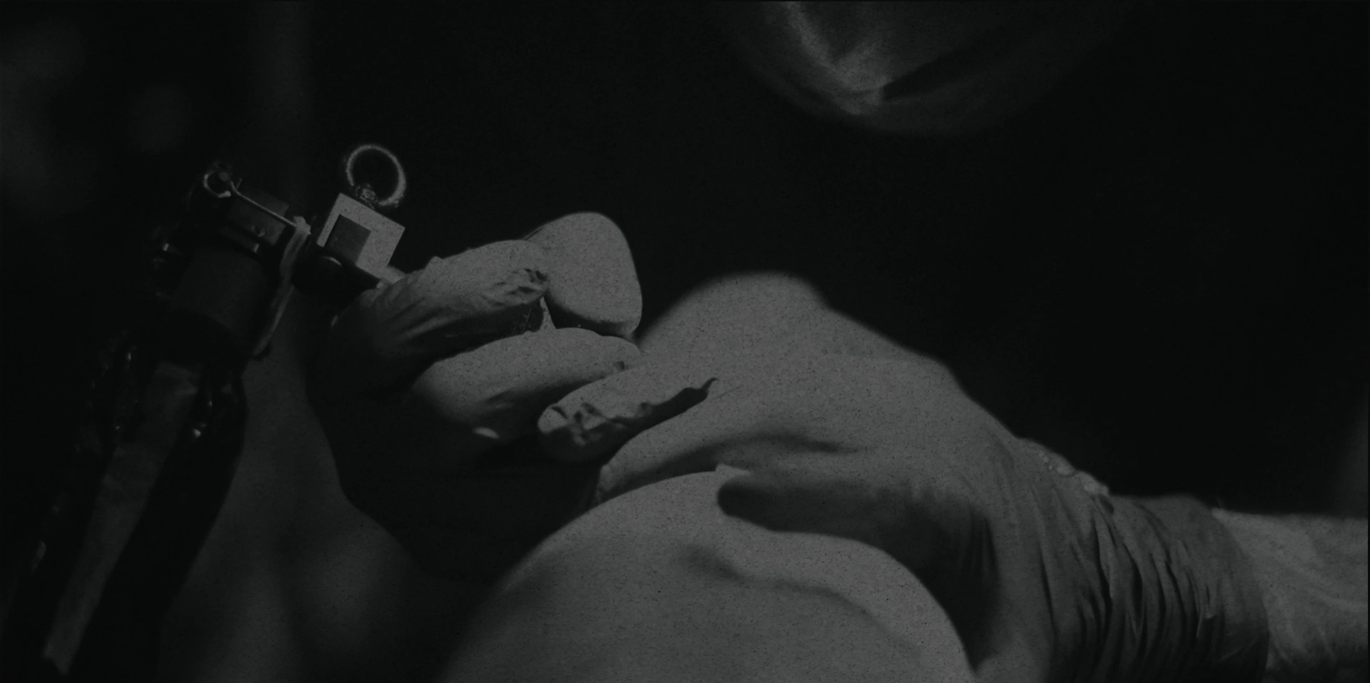 A close-up, black and white image of a gloved hand holding a tattoo machine at new spider ink, a tattoo studio in Dublin. The scene is dimly lit, creating a dramatic and focused atmosphere as ink meets skin.